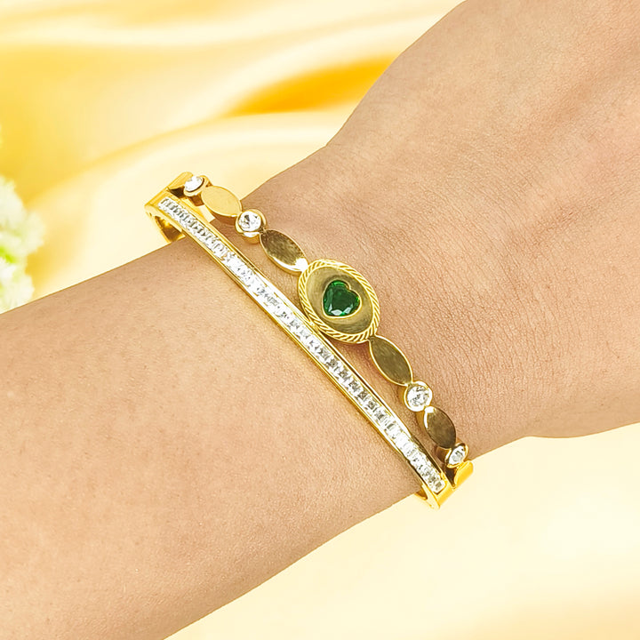 A gold bangle bracelet with clear crystals and a heart-shaped green gemstone on a wrist.