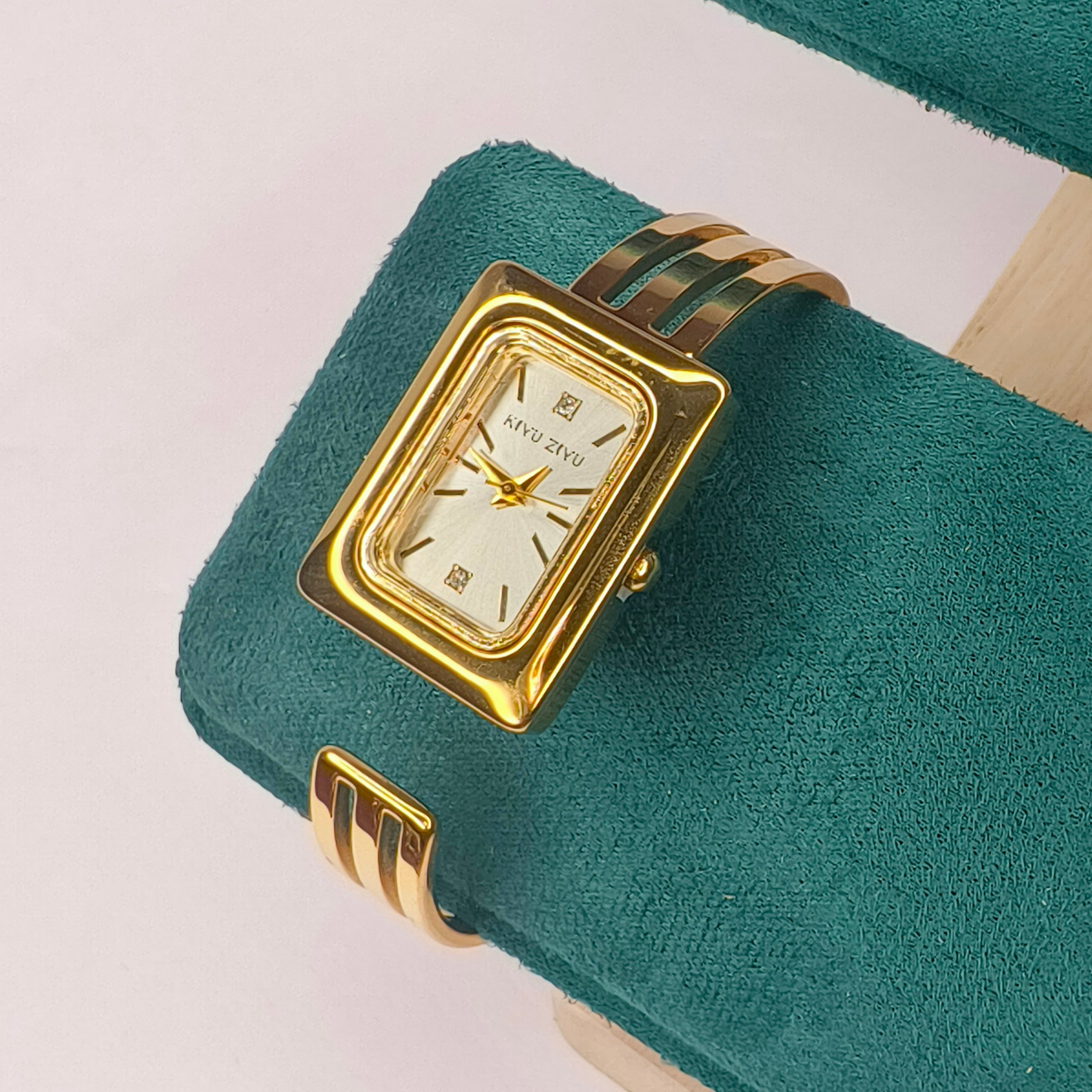 Elegant gold rectangular women's watch with a metallic band on a teal velvet cushion.